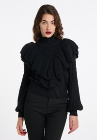 Schwarzer Strickpullover mit hohem Kragen und gestuften Rüschendetails. Verfügt über eine Zopfmusterstruktur und ausgestellte Ärmel. Getragen mit eng anliegenden schwarzen Hosen.