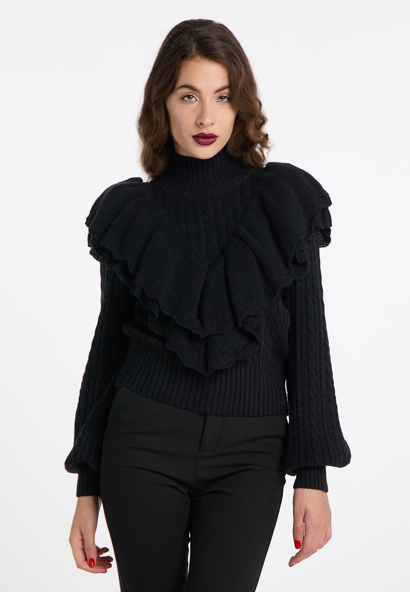 Schwarzer Strickpullover mit hohem Kragen und gestuften Rüschendetails. Verfügt über eine Zopfmusterstruktur und ausgestellte Ärmel. Getragen mit eng anliegenden schwarzen Hosen.