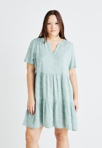 Short-sleeve, light green dress with a V-neck, gathered waist, and a tiered ruffle hem, featuring a subtle white leaf pattern throughout.