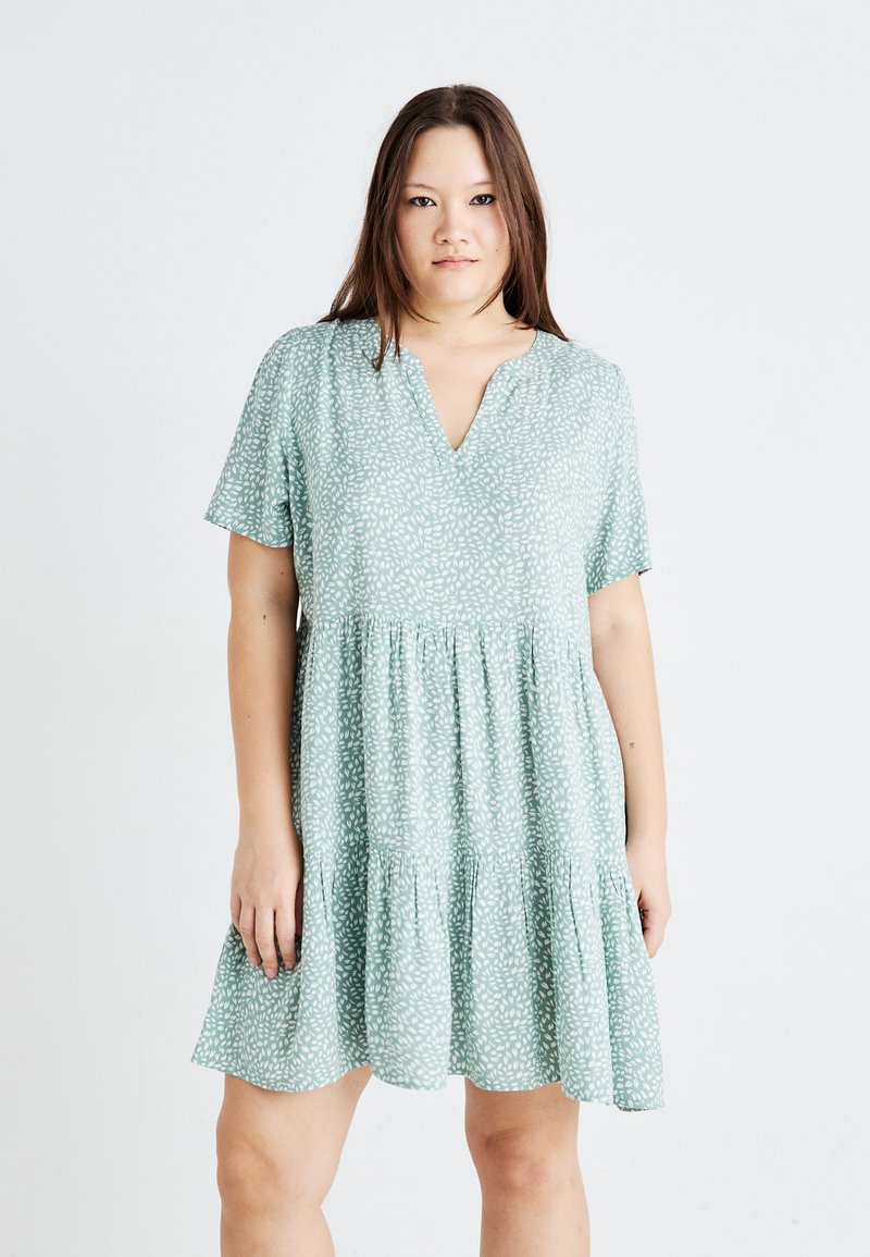 Vestido de manga corta, color verde claro, con escote en V, cintura fruncida y un dobladillo de volantes en capas, que presenta un sutil patrón de hojas blancas a lo largo.