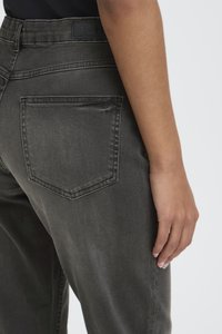 Prawa strona osoby noszącej ciemnoszare dżinsy z widoczną tylną kieszenią i rozluźnioną ręką na jasnym tle.