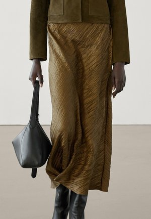 Personne portant une jupe midi marron texturée, une veste vert olive, des bottes noires montantes jusqu'aux genoux, et tenant un sac à main en cuir noir.