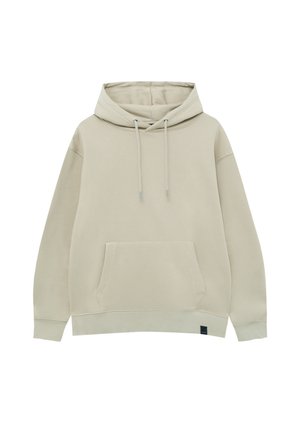 Personne portant un sweat à capuche beige, un jean large bleu clair et des baskets blanches avec des semelles marron, debout devant un fond uni.