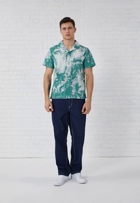 Camisa de manga corta verde y blanca con un diseño tie-dye y cuello, combinada con jeans azul oscuro. Zapatillas blancas completan el atuendo.