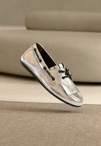 Μεταλλικό ασημί παπούτσι τύπου slip-on με μαύρα κορδόνια, με υφή στην επιφάνεια και επίπεδη σόλα, παρουσιασμένο σε ουδέτερο φόντο.