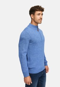 Mand iført en blå halvmønstret sweater og mørke jeans, stående sidelæns mod en ensfarvet hvid baggrund.