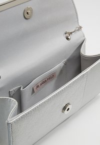 Portafoglio argento con un esterno texturizzato, chiusura a scatto e scomparti interni. Presenta un'etichetta con la scritta "ANNA FIELD" con una foderatura liscia.