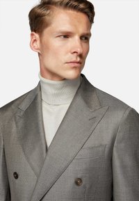 Double-breasted grey blazer with textured fabric, notched lapels, and two brown buttons, worn over a light turtleneck sweater.