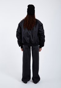 Black bomber jacket with quilted sleeves and a ribbed hem, paired with high-waisted black wide-leg jeans. Knitted black beanie on head.