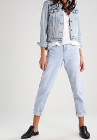 Light blue denim jacket with two chest pockets, paired with rolled-up light blue jeans and black slip-on shoes. Simple white top underneath.