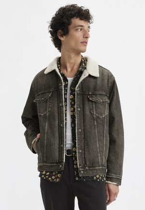 Donkere grijze spijkerjack met een zachte, crèmekleurige schapenvachtkraag. Voorzien van twee borstzakken en zichtbare stikseldetails. Zwarte shirt eronder.