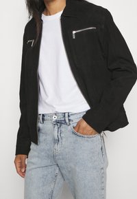 Svart mockajacka med dragkedja fram, två framfickor med dragkedja och en avslappnad passform. Kombinerad med ljusblå jeans och en vit t-shirt.