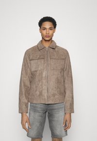 Giacca in faux suede marrone con zip frontale, colletto e due tasche sul petto. Indossata con pantaloni corti grigi, caratterizzati da un design semplice e testurizzato.
