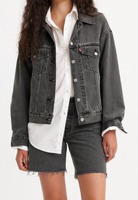 Chaqueta de mezclilla negra con dos bolsillos en el pecho, botones plateados y un corte relajado. Llevada sobre una camisa blanca de botones y combinada con pantalones cortos negros.