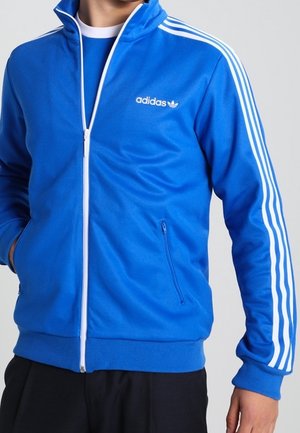 Uomo che indossa una giacca Adidas blu con cerniera, strisce bianche sulle maniche e logo Adidas sul lato sinistro del petto, con le mani nelle tasche.