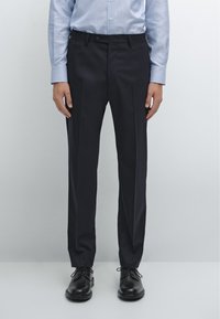 Marineblauwe broek met voorplooien, een gladde textuur en een slim fit. G gedragen met een lichtblauw gestreept overhemd en zwarte veterschoenen.