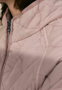 Veste matelassée rose avec une fermeture éclair, des motifs de couture en relief et une texture douce, mettant en valeur le design du col arrondi.
