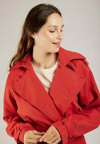 Manteau trench rouge avec un large col, des manches longues et une ceinture. Caractéristiques d'un tissu texturé et de fermetures à boutons, porté par-dessus un haut léger.