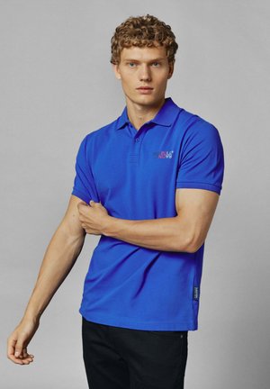 Krótki rękaw, gładka koszulka polo w królewskim niebieskim kolorze, z dwiema guzikami przy kołnierzyku oraz małym logo "RED BULL" na piersi.