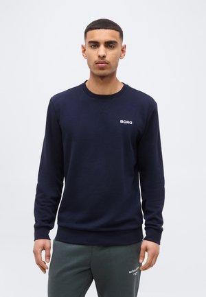 Uomo con felpa blu scuro e pantaloni sportivi grigi, in piedi davanti a uno sfondo chiaro e uniforme, rivolto verso la fotocamera.