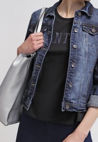 Farkkutakki, jossa on metalliset napit, puettuna mustan T-paidan päällä. Hopea kassi, jossa on teksturoitu pinta. Käsi sijoitettu kassin hihnalle.