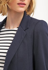 Marindblå blazer med strukturerade axlar, tillverkad av textil med struktur, ihop med en svart och vit randig topp. Närbild.