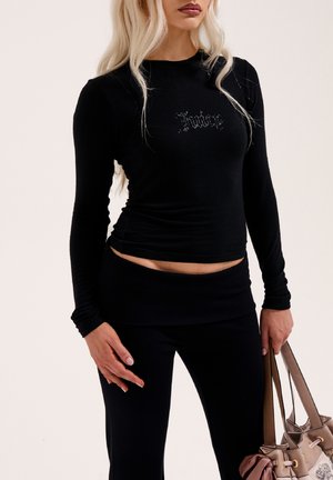 Top nero a maniche lunghe con design aderente e accento di strass "Khadese", abbinato a leggings neri e una borsa di colore chiaro.