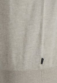 Suéter de punto beige claro con una textura suave, dobladillo acanalado y una pequeña etiqueta negra en el lateral. Diseño sencillo con detalles mínimos.