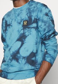 Cotton sweatshirt in tie-dye blue with darker blue swirls. Features ribbed cuffs and hem, and a small rectangular logo patch on the chest.