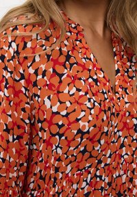 Hedvábná blůza s červeným a oranžovým vzorem, s volánovým výstřihem, obsahující nepravidelné kruhové tvary na námořnickém pozadí a texturovanou látkou.