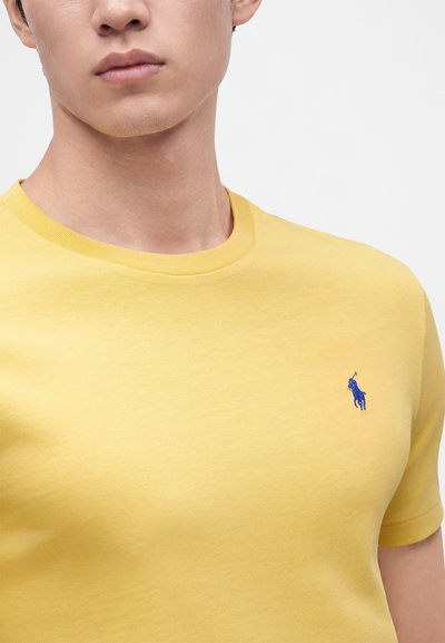 Νεαρός άνδρας που φοράει απλό κίτρινο t-shirt με λαιμόκοψη και μικρό μπλε λογότυπο παίκτη πόλο στο αριστερό στήθος.