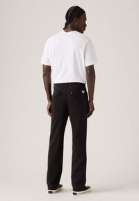 Zwarte broek met een rechte pijp en twee achterzakken, gecombineerd met een witte t-shirt met korte mouwen. De broek heeft een klein rood labeldetail.