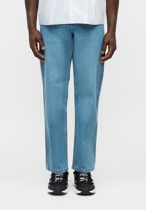 Homme portant un jean droit bleu clair avec le texte "Versace Jeans Couture" sur les coutures extérieures, des baskets noires et blanches, et une chemise blanche.