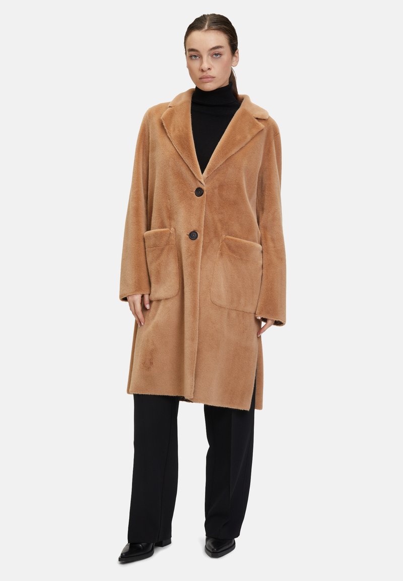 Cappotto in misto lana color beige con ampio colletto, due tasche frontali a toppa e chiusura con bottoni neri. Il modello indossa un dolcevita nero e pantaloni.