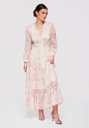 Vrouw in een lange witte jurk met roze bloemenprint, kanten details, lange pofmouwen en roze hoge hakken, poserend tegen een effen achtergrond.