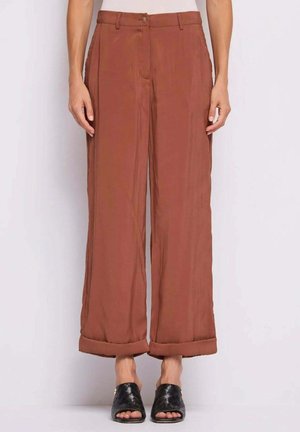 Donna che indossa pantaloni a gamba larga color ruggine con orli risvoltati e scarpe nere intrecciate aperte sulle dita, in piedi contro uno sfondo semplice.