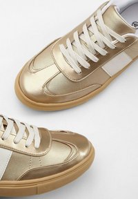 Sneaker metallici dorati con accenti bianchi, dotati di una texture liscia, punta rotonda e suola in gomma piatta, allacciati con stringhe bianche.
