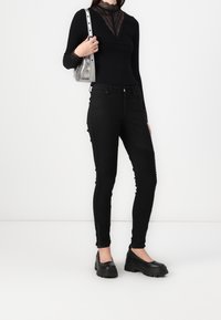 Jeans neri aderenti con dettagli rigati sulle cosce, abbinati a un top nero a maniche lunghe con applicazioni in pizzo e una borsa a spalla argento.
