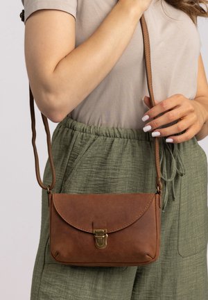 Sac en cuir brun porté en bandoulière avec un rabat arrondi, fermeture à boucle en laiton et une sangle réglable, tenu par une personne portant une chemise claire et un pantalon vert.