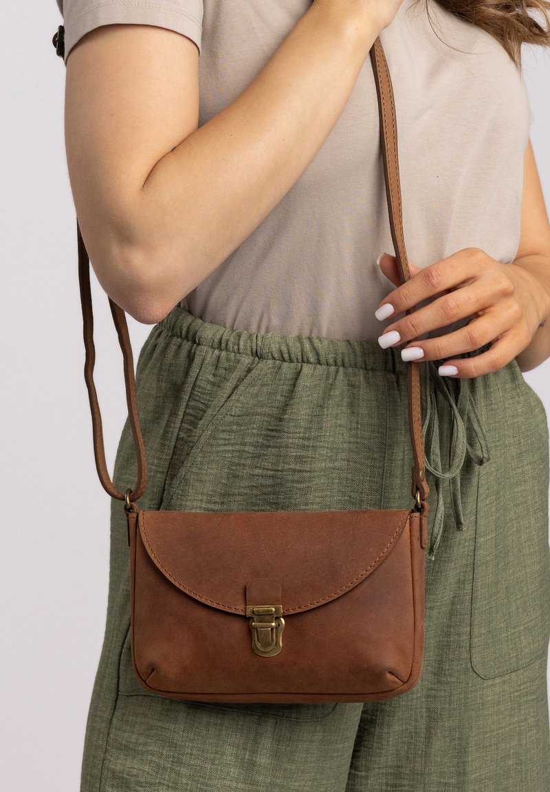 Žena oblečená v béžovom topi a zelených nohaviciach, ktorá drží hnedú koženú tašku cez rameno so mosadzným západkou za popruh.