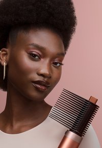 Hairstylingborstel met een zwart en koperen ontwerp, voorzien van fijne tanden en een gestructureerde grip, gehouden tegen een roze achtergrond.