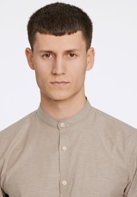 Μπεζ πουκάμισο με κούμπωμα και γιακά μανδαρίνου, κατασκευασμένο από λείο ύφασμα με διακριτική υφή, με τρία κουμπιά στο μπροστινό μέρος.