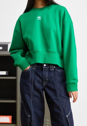 Person iført grøn Adidas-sweatshirt og mørkeblå denimbukser med hvide syninger, hånd i lommen, stående indenfor.