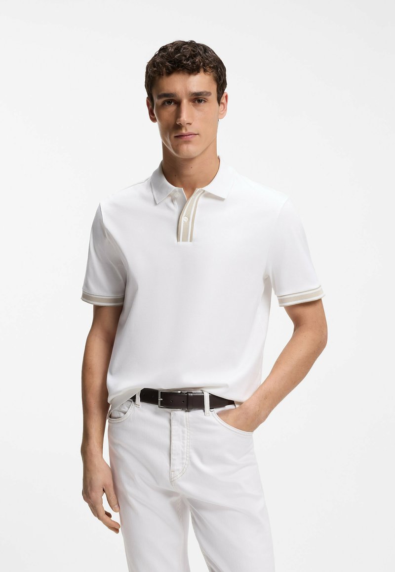 Homme portant un polo blanc à manches courtes avec un bord beige sur le col et les manches, un pantalon blanc, une ceinture foncée, debout avec une main dans la poche.