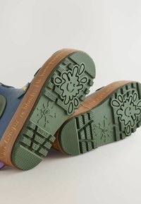 Bunte wasserdichte Stiefel mit einem blauen Obermaterial, grüner Gummisohle und strukturiertem Profil, das verspielte Designs und erhabene Details aufweist.