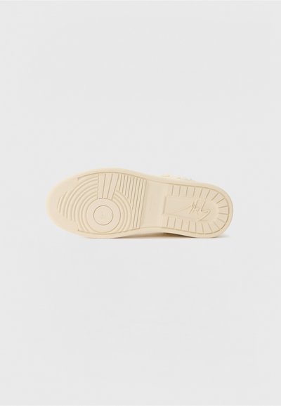 Cremfarvet sneakersål med en tekstureret gummioverflade, der har cirkulære og lineære mønstre. Markeringer og et logo er til stede på sålen.