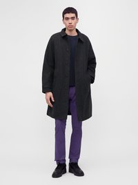 Young man wearing a dark buttoned coat, black shirt, purple pants, and black shoes standing against a plain light background.