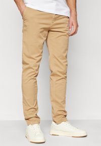 Chino beige in cotone con una vestibilità slim, dotati di tasche laterali e una texture liscia, abbinati a sneakers bianche.