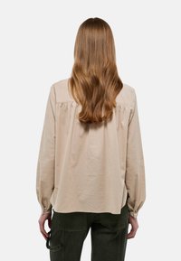 Blouse beige à manches longues avec une coupe décontractée, détail froncé au dos et poignets boutonnés, associée à un pantalon vert foncé. Texture en coton doux.