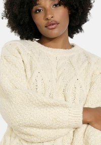 Svetre krémovej farby s pletenými vzormi kabeliek, okrúhlym výstrihom a rebrovanými manžetami. Textúrovaná, hrubá priadza prispieva ku pohodlnému vzhľadu.